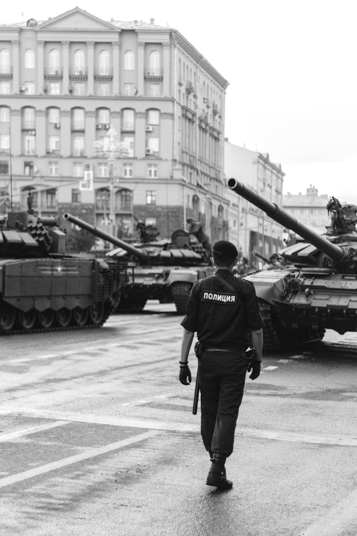 my-steps Monochrome image of a police officer walking amidst military tanks in an urban setting.